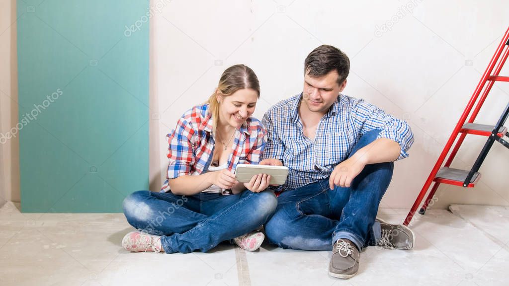 Smiling young couple sitting on floor at new house under construciton and browsing internet