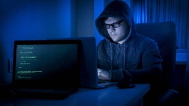 Karanlık odada oturan ve dizüstü bilgisayarda çalışan genç hacker portresi