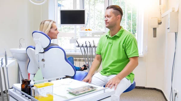 Stilig hane tandläkare talar till hans ung kvinna patient i tandläkare kontor — Stockfoto