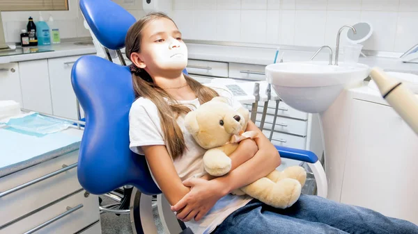 Cute girl with teddy bear sitting in dentist chair in clinic and feeling nervous before treating teeth — Stock Photo, Image