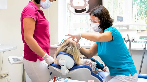 Dentista em máscara portativa e óculos treting paciente do sexo feminino sentado na cadeira — Fotografia de Stock