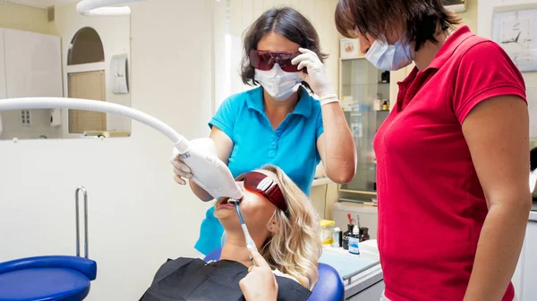 Dentista e assistente preparando jovem mulher sentada na cadeira do dentista para o procedimento de clareamento dos dentes — Fotografia de Stock