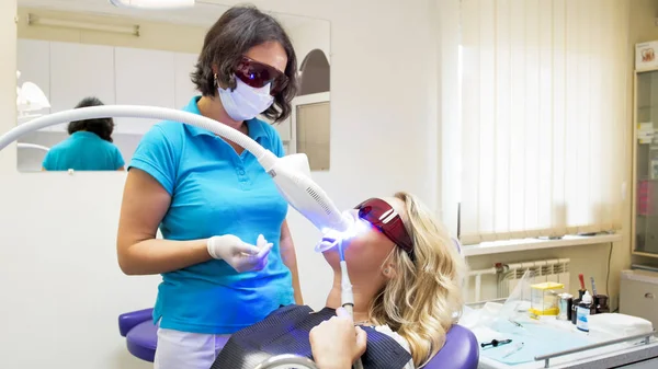 Tandarts in beschermende oranje bril permanent naast vrouw tijdens tanden whitening procedure — Stockfoto