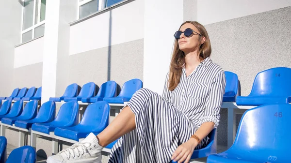 Sexy stylish young woman in sunglasses sitting on empty stadium seats in bright sunny day — Stock Photo, Image