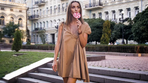 Hermosa joven en vestido posando en la calle y lamiendo piruleta — Foto de Stock