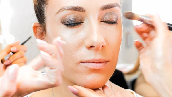 Closeup portrait of young model sitting with eyes closed while makeup artist applies makeup on her face — Stock Photo, Image