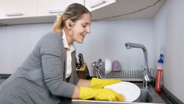 Imágenes de 4k de la feliz mujer sonriente escuchando música con auriculares mientras lava los platos en la cocina — Vídeos de Stock