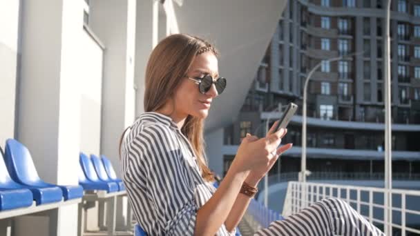 Primer plano 4k metraje de la hermosa joven sentada en la calle en el día soleado y el uso de teléfono inteligente — Vídeos de Stock