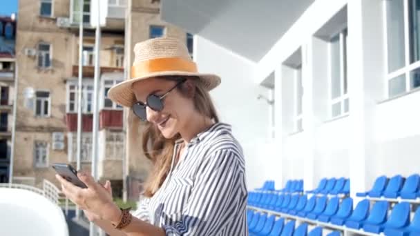 4k video de la joven feliz en gafas de sol y sombrero usando amrtphone en la calle en el día soleado — Vídeos de Stock