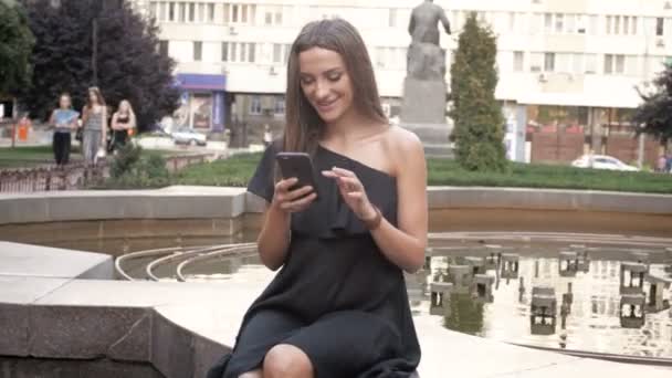 Mulher bonita em vestido preto sentado no parque e usando smartphone — Vídeo de Stock