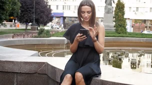 4k кадри красивої елегантної жінки в чорній сукні, що сидить у парку фонтанів і використовує смартфон — стокове відео