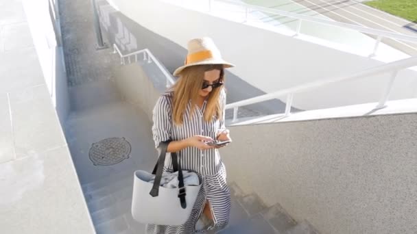 Zeitlupenvideo einer jungen Frau mit Hut und Sonnenbrille, die mit ihrem Smartphone die Treppe auf der Straße hinaufläuft — Stockvideo