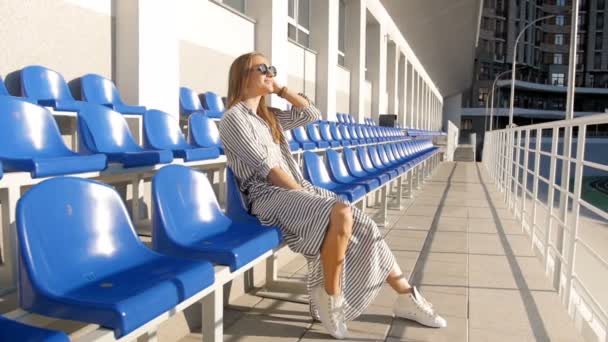 Vidéo au ralenti de femme élégante en lunettes de soleil et robe longue posant sur arène vide — Video