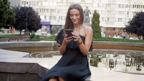 Vidéo au ralenti de belle jeune femme assise à la fontaine et utilisant un smartphone — Video