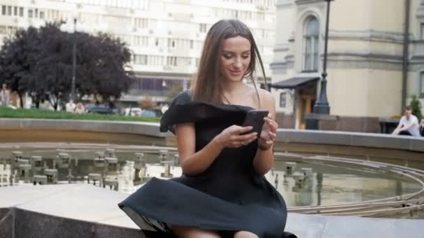 Filmagem em câmera lenta de jovem sexy em vestido curto sentado no parque no dia ventoso e digitando mensagem no smartphone — Vídeo de Stock
