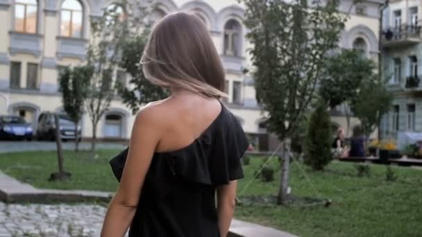 Vídeo en cámara lenta de la hermosa mujer sonriente en vestido negro caminando en el parque y mirando a la cámara — Vídeos de Stock