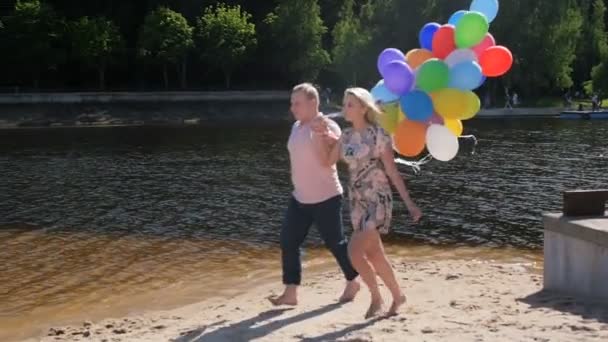 Vidéo au ralenti d'un jeune couple heureux tenant un gros tas de ballons et courant sur la plage — Video