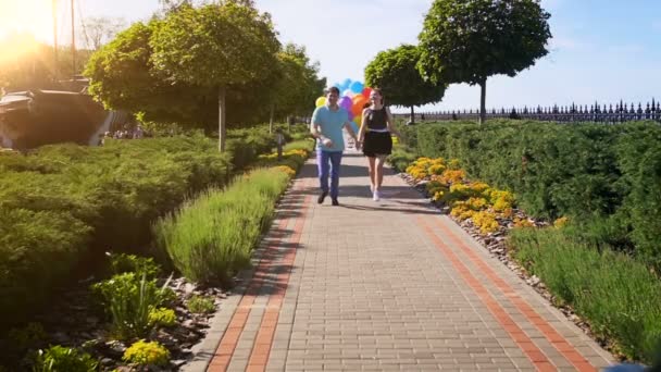 Vídeo en cámara lenta de la feliz pareja riendo sosteniendo un gran montón de globos y corriendo en el parque — Vídeo de stock