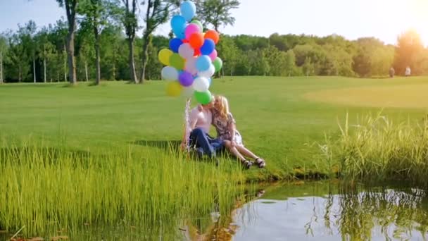 Vídeo en cámara lenta de una hermosa pareja sosteniendo globos de colores y sentada en la orilla del río — Vídeo de stock