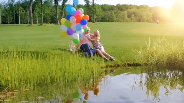 Vídeo en cámara lenta de feliz pareja joven enamorada sosteniendo globos de colores y relajándose junto al río — Vídeo de stock