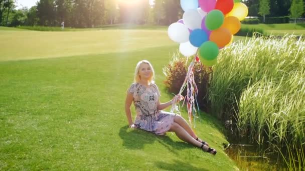 Slow motion beelden van mooie blonde meisje met kleurrijke ballonnen, zittend op het gras — Stockvideo