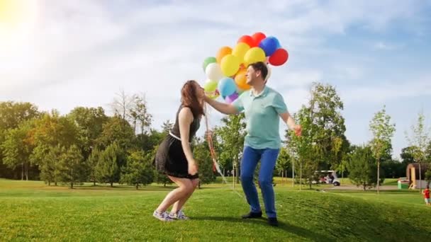 Imágenes en cámara lenta de feliz pareja joven sosteniendo un montón de globos de colores y saltando en el aire — Vídeos de Stock