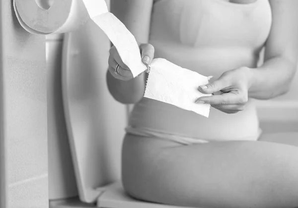 Black and white image of young woman sitting on toilet and tearing off toilet paper — Stock Photo, Image