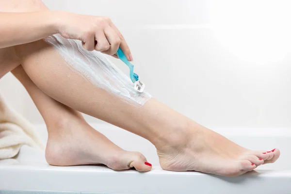 Imagen de cerca de una joven sentada en el baño y depilando las piernas con una navaja de afeitar — Foto de Stock