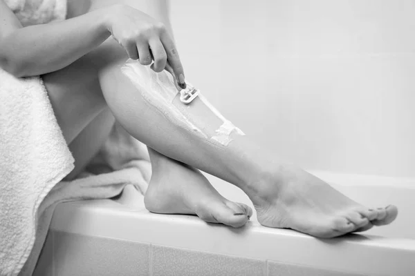 Black and white image of young woman sitting in bathroom and shaving legs — Stock Photo, Image