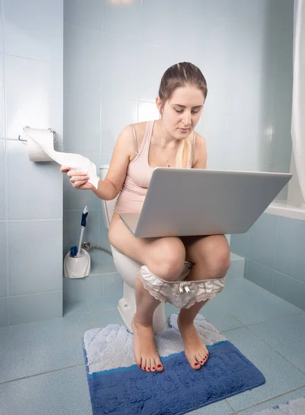 शौचालय पर बैठे हुए लैपटॉप का उपयोग करने वाली युवा ब्रुनेट महिला — स्टॉक फ़ोटो, इमेज
