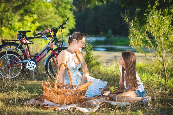Feliz madre joven haciendo un picnic bajo el árbol cerca del río con su hija — Foto de Stock