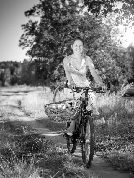 Black and white image of beautiful smiling woman riding on bicycle with big basket for picnic in park — Stock Photo, Image