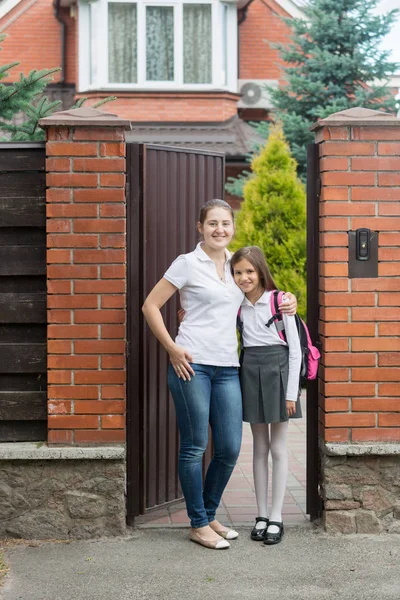 Jovem mãe feliz posando com a filha em uniforme escolar na frente da casa — Fotografia de Stock