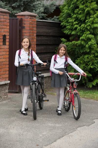 Две улыбающиеся девочки со школьными сумками ходят на велосипедах по улице — стоковое фото