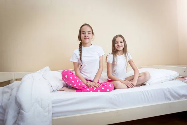 Duas irmãs sorridentes de pijama sentadas na cama e olhando para a câmera — Fotografia de Stock