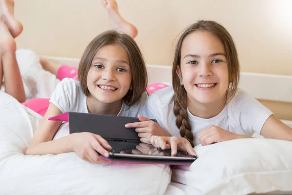 Portrait of two smiling girls lying on bed with digital tablet and looking in camera — Stock Photo, Image