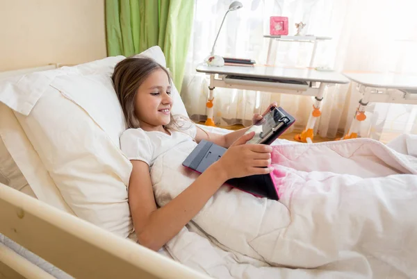 Портрет улыбающейся девочки-подростка, лежащей в постели и просматривающей интернет на цифровом планшете — стоковое фото