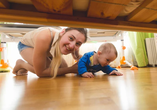 Piękna młoda matka z toddler chłopiec leży na podłodze i patrząc pod łóżkiem — Zdjęcie stockowe