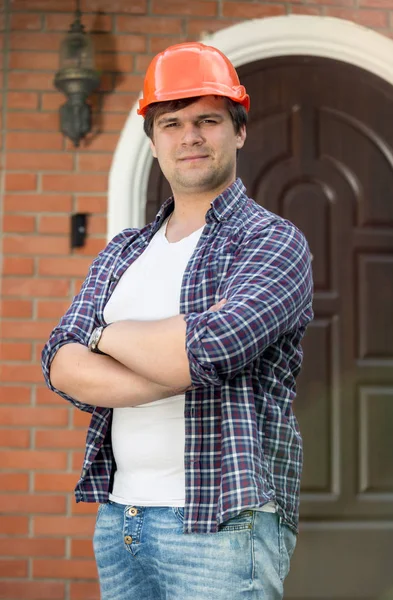 Portrait of handsome young man in checkered shirt and hardhat posing in front of house doors — Stock Photo, Image