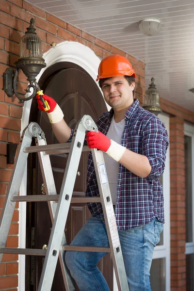 Retrato de electricista sonriente en casco protector de plástico de pie sobre escalera —  Fotos de Stock