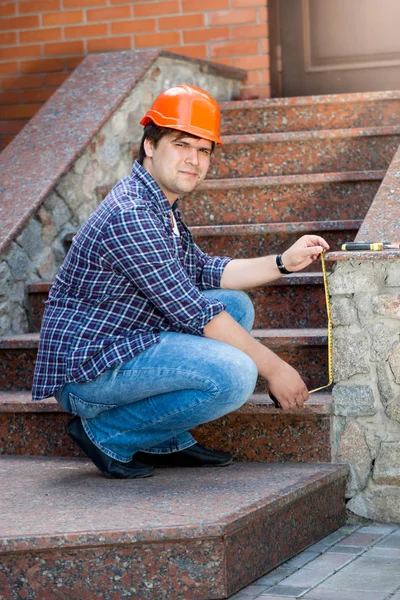 Young builder in hardhat measuring stone stairs in house with measuring tape — Stock Photo, Image