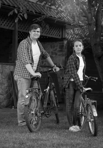 Black and white photo of young man with teenage daughter posing with bicycles at house backyard — Stock Photo, Image