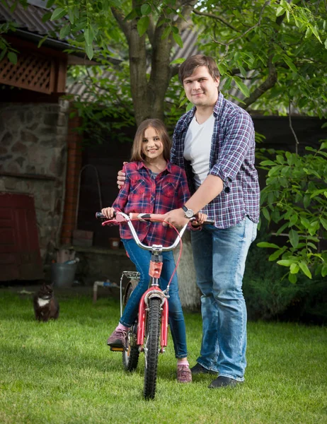 Sonriente joven enseñando a su hija a montar en bicicleta en el patio trasero de la casa — Foto de Stock