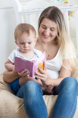 Onun bebek çocukla puf üzerinde oturan ve ona kitap okuma gülümseyen genç anne portresi