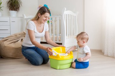 Beautiful young mother sitting on floor with her baby son and teaching him using toilet pot clipart