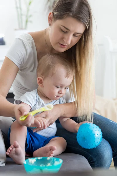 Joven madre sentada en el sofá con su hijo bebé y alimentándolo con cuchara — Foto de Stock