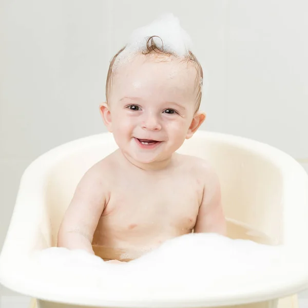 Retrato de bebê sorridente bonito menino com espuma de sabão na cabeça sentado no banho e olhando para a câmera — Fotografia de Stock