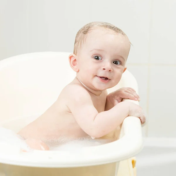 Carino sorridente bambino ragazzo seduto in bagno con schiuma e guardando in fotocamera — Foto Stock