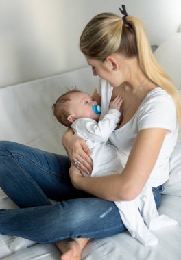 Yatağın üstüne oturan ve uyuyan bebeği tutan güzel genç kadın