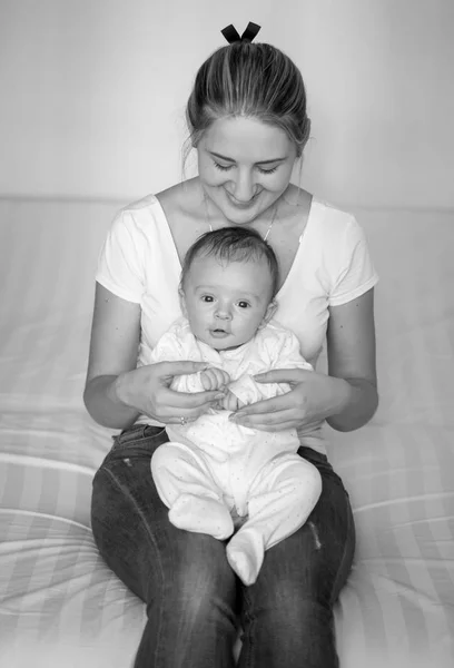 Foto em preto e branco de sorrir jovem mãe sentada na cama e segurando seu bebê — Fotografia de Stock